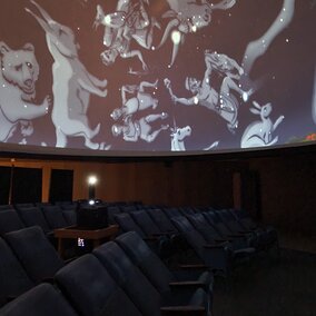 Image inside of the Mueller Planetarium showing images of the constellations, the projector in the middle of the room, and the chairs.